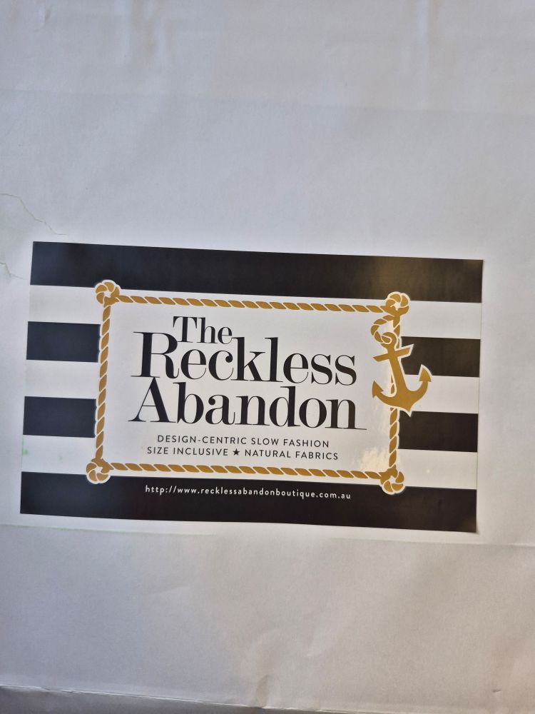 A sticker label on a white paper bag labelled "The Reckless Abandon" with an anchor and rope border, and a background of thick horizontal black and white stripes. Subtext saying "design centric slow fashion • size inclusive • natural fabrics" and the website below, "http://www.recklessabandonboutique.com.au".
