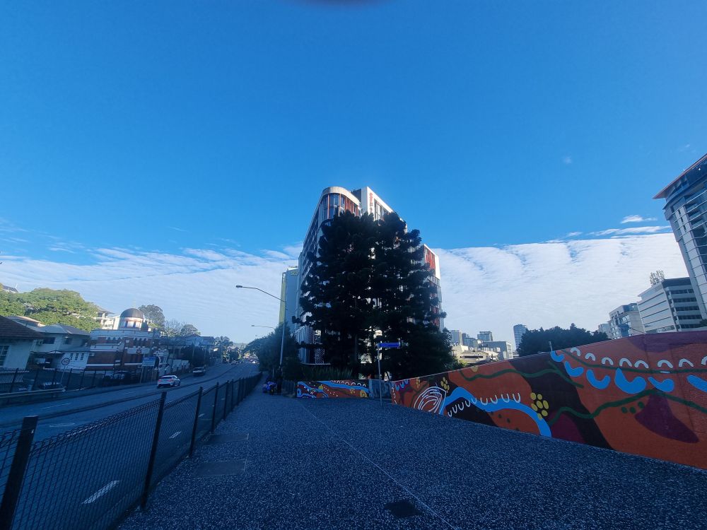 A view down a shadowed street, contrasting the blue sky containing a stark straight division of light, white clouds filling the bottom half of it in a front. There's a wall featuring bold patterned artwork in oranges and blue.