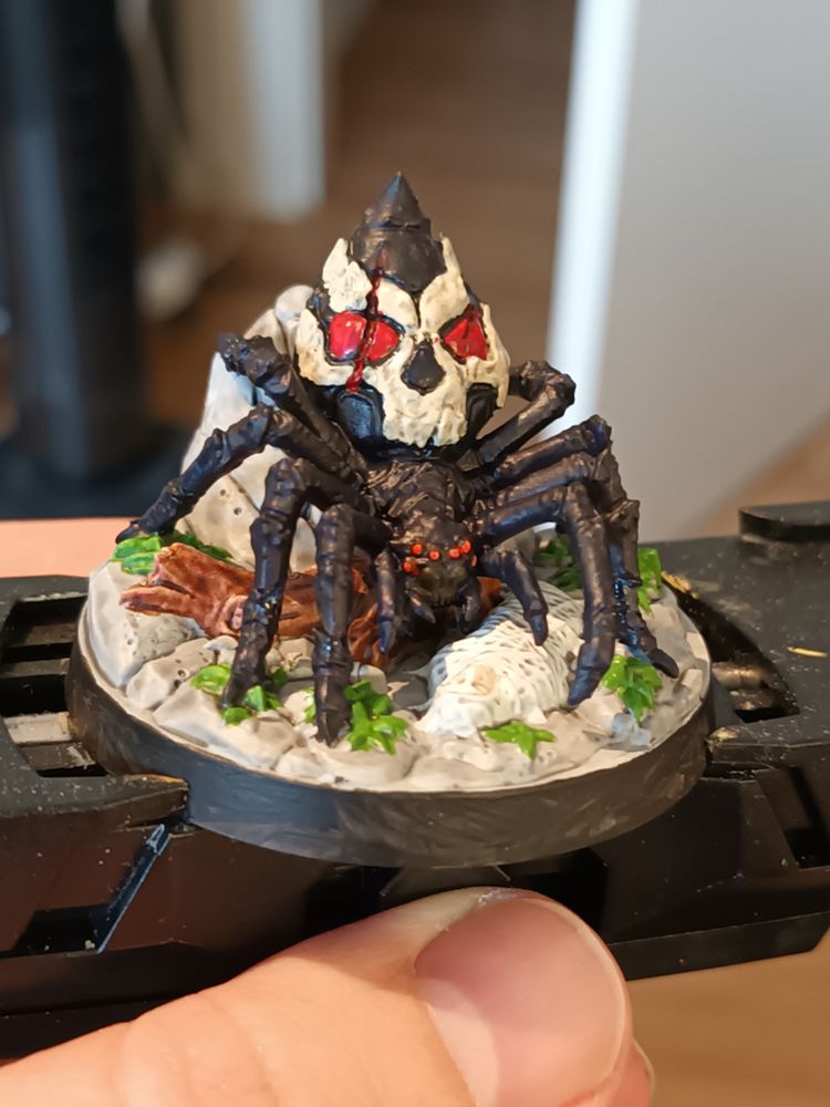 A giant spider on a rock with a webbed-up human below it, next to a fallen tree. The Spider has a Skull pattern marred by gashes in its exoskeleton
