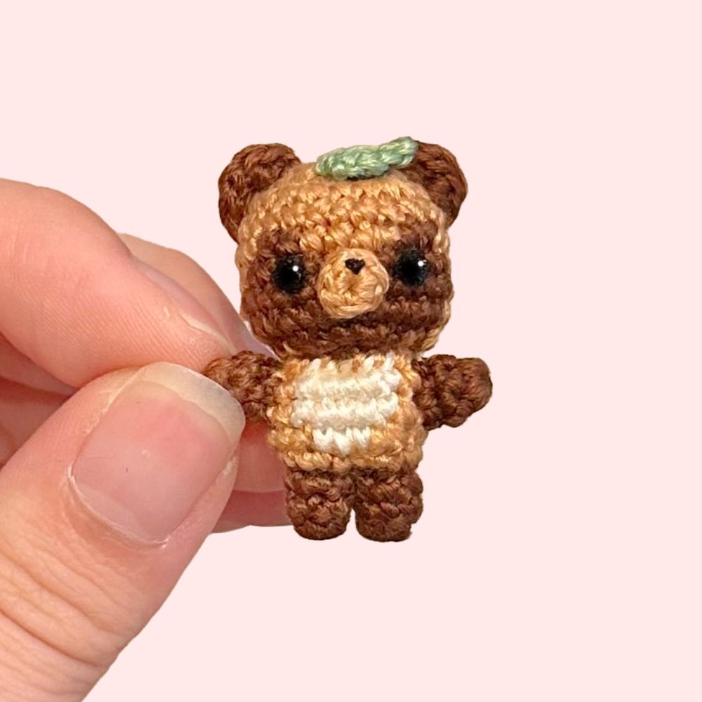 Microcrochet of a tanuki (raccoon-dog) in dark brown, brown, and white, with a single pastel green leaf on its head. The tanuki is held up by one hand on the left, on a pink background
