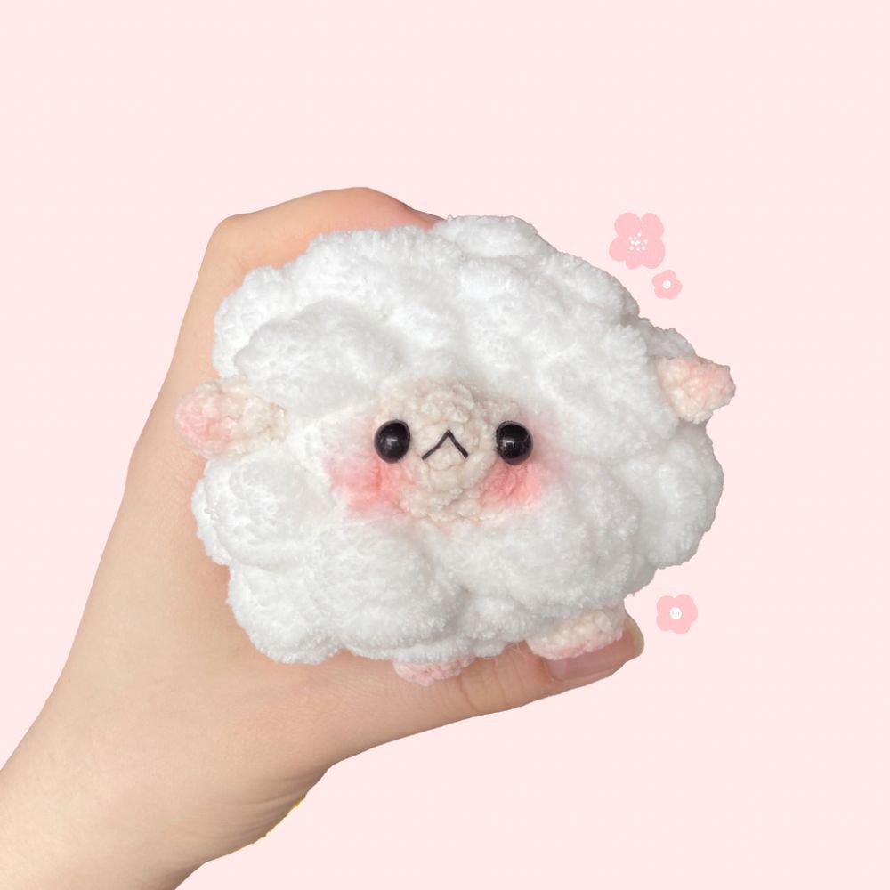 Hand holding up a crochet sheep shaped like a cloud, with a small face, ears, and legs poking out from the main body. Blush is added to the cheeks and ears