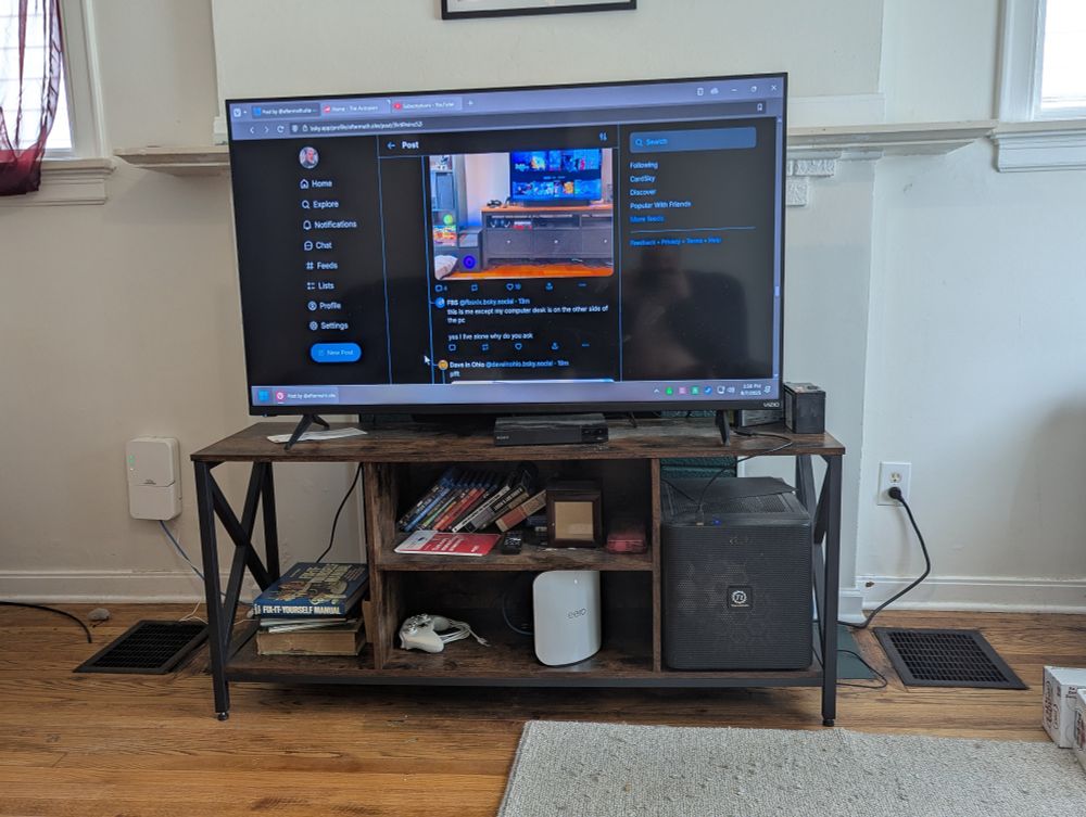 A 55 inch TV on a stand. The left part of the stand has random books inside it. The middle sections have Blu-rays and various computer-related things. The right side is fully taken up by a large square computer case.