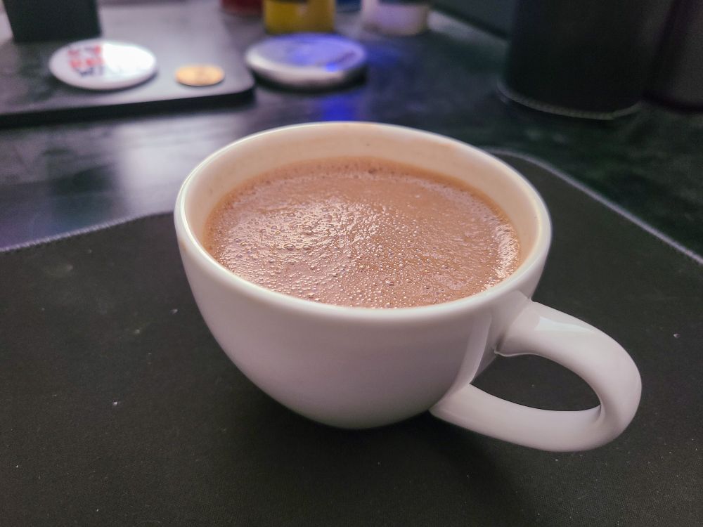 A small, white mug with a handle filled with frothy hot chocolate on a black desk mat. In the background, on a dusty desk, are a Harris-Walz campaign button, an American one-dollar coin, a metal perpetual calendar, and a pen holder.