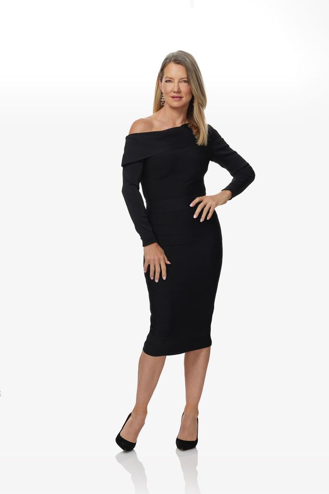 A blonde haired woman in a black dress and heels is posing with a white background behind her. 