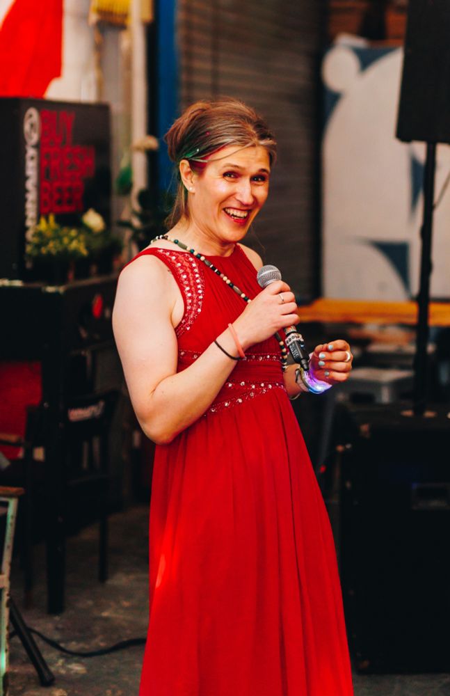 Picture shows a woman wearing a red dress holding a microphone and laughing