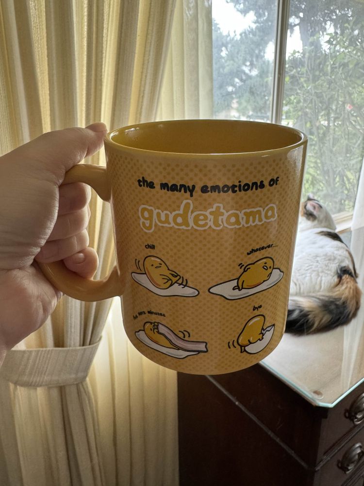 white hand holding a yellow mug that has Gudetama all over it, and in the background is a calico cat on a buffet looking out a window 