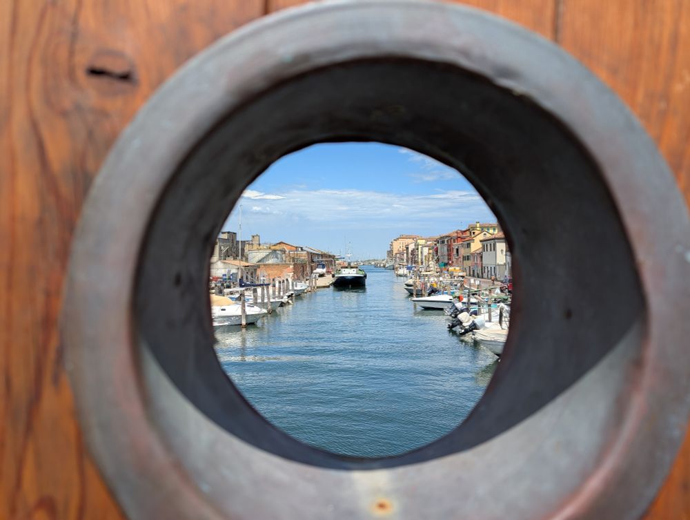 Blick durch ein großes Loch aus Metall in einem Holzzaun, dahinter ein Kanal gesäumt von Booten, Anlegestegen und italienischen Häusern. 