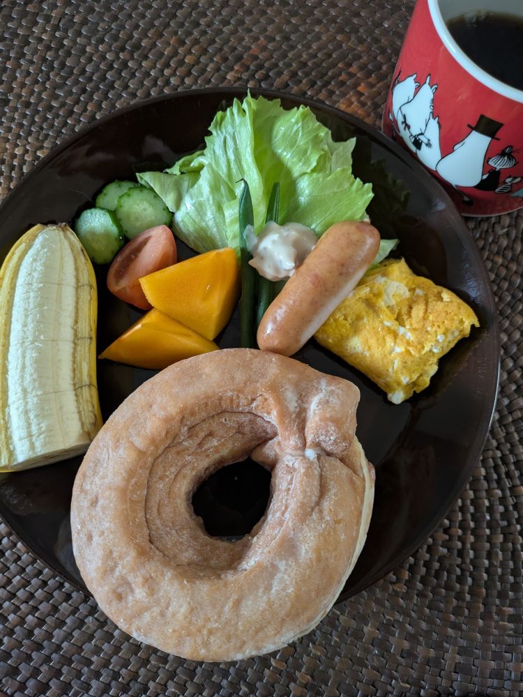 赤いコーヒーカップ、濃い茶色の皿に生ドーナツ、玉子焼き、ウインナ、インゲン豆、レタス、きうり、トマト、柿、バナナ