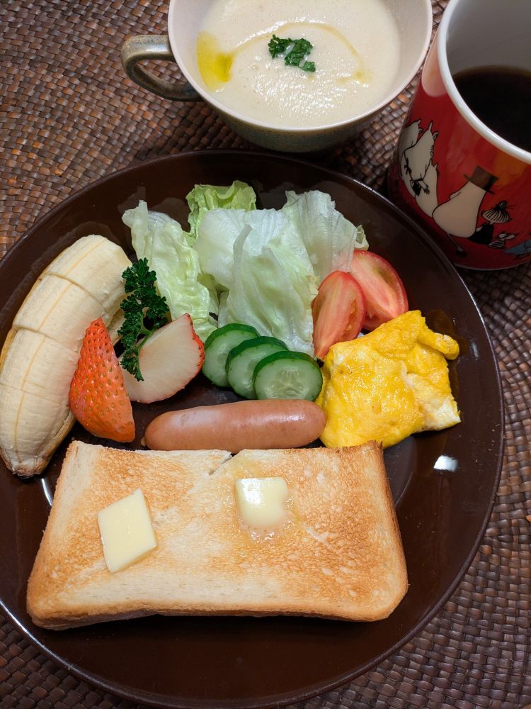 大根ポタージュスープのカップ、赤いコーヒーカップ、濃い茶色の皿にバタートースト、玉子焼き、トマト、レタス、きうり、ウインナ、イチゴ、バナナ