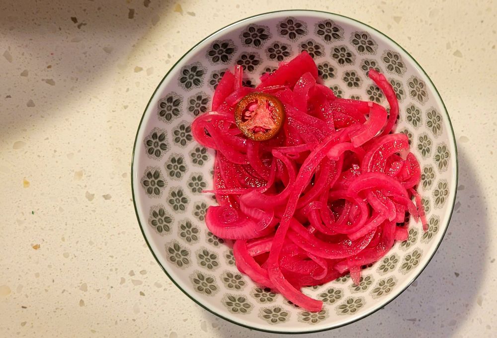A green bowl sits on a white counter. Inside it lies a pile of neon pink pickled onions and one slightly pink slice of serrano