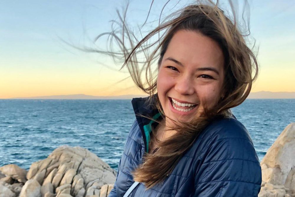 A portrait of Kendra Chan standing on the coast, laughing as the wind blows her hair.