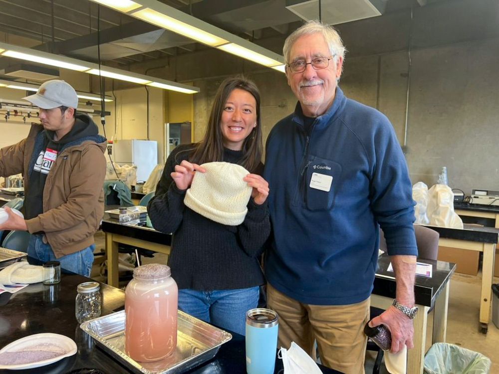 Two people standing at a lab bench, creating dye from purple urchin spines. One is holding up a wool beanie that they will color with the natural dye.