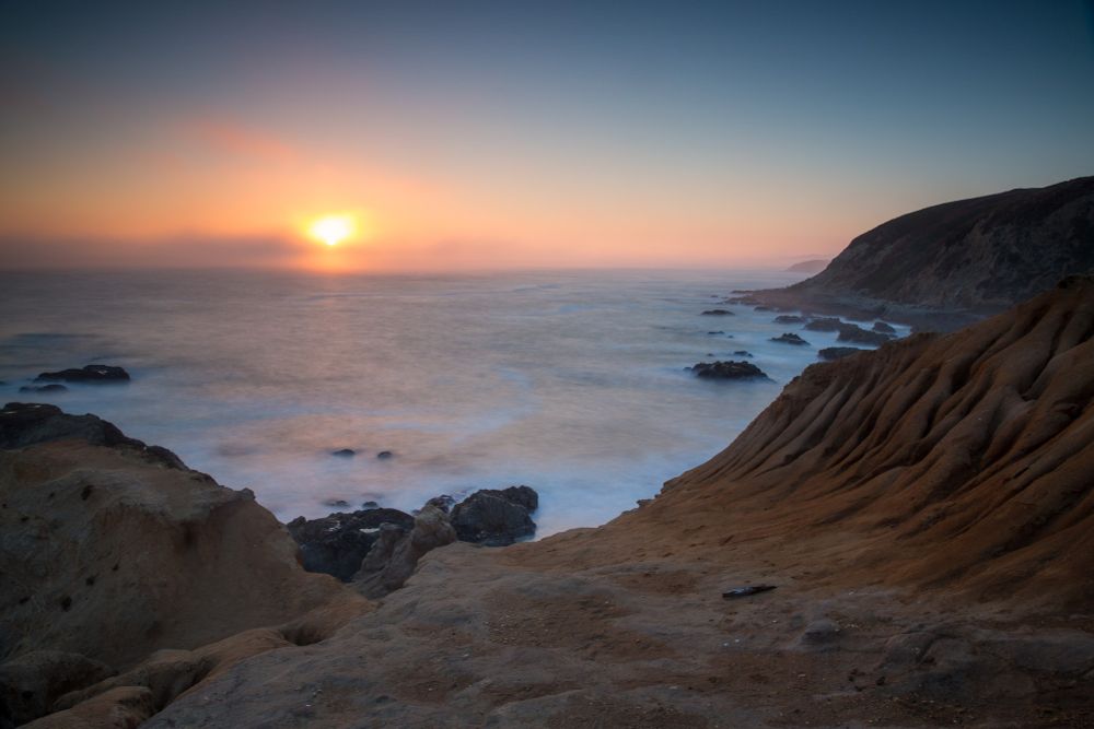 The sun rising over the sea, seen from a rocky coastal bluff.