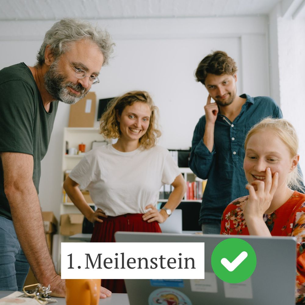 Ein Team aus vier Personen steht in einem Büro und schaut gemeinsam auf einen Laptop, dabei lächeln sie. Auf dem Bild befindet sich eine Kachel mit dem Text „1. Meilenstein“ und einem weißen Haken in einem grünen Kreis.
