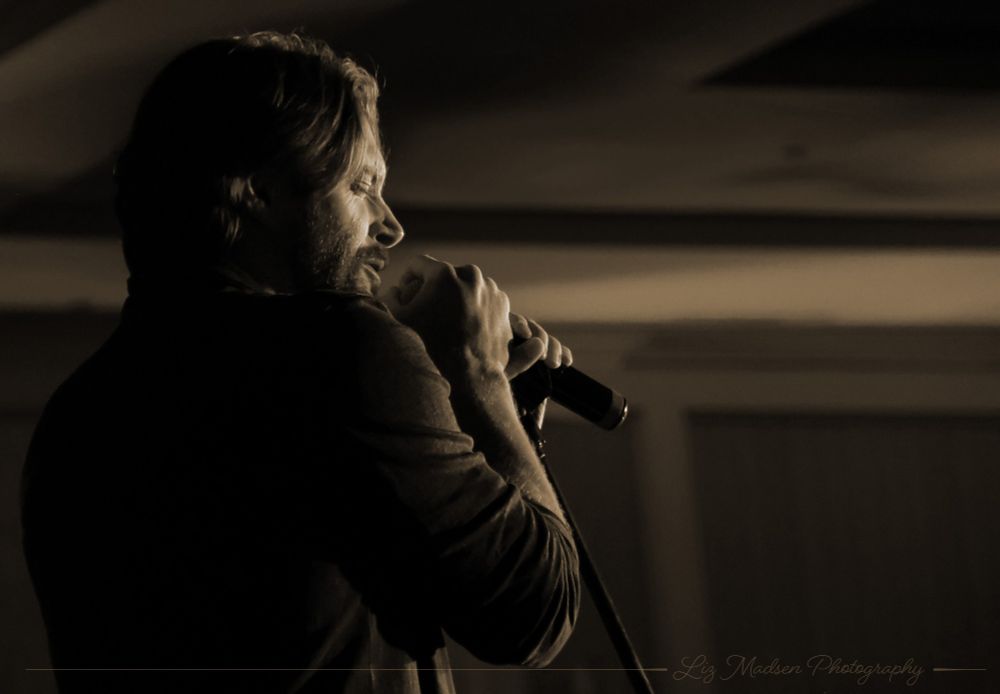 A sepia-toned photo of Jensen singing at the DC convention. The camera looks over his right shoulder & we see his face in profile. He has both hands on the microphone & his eyes are closed as he feels the music.