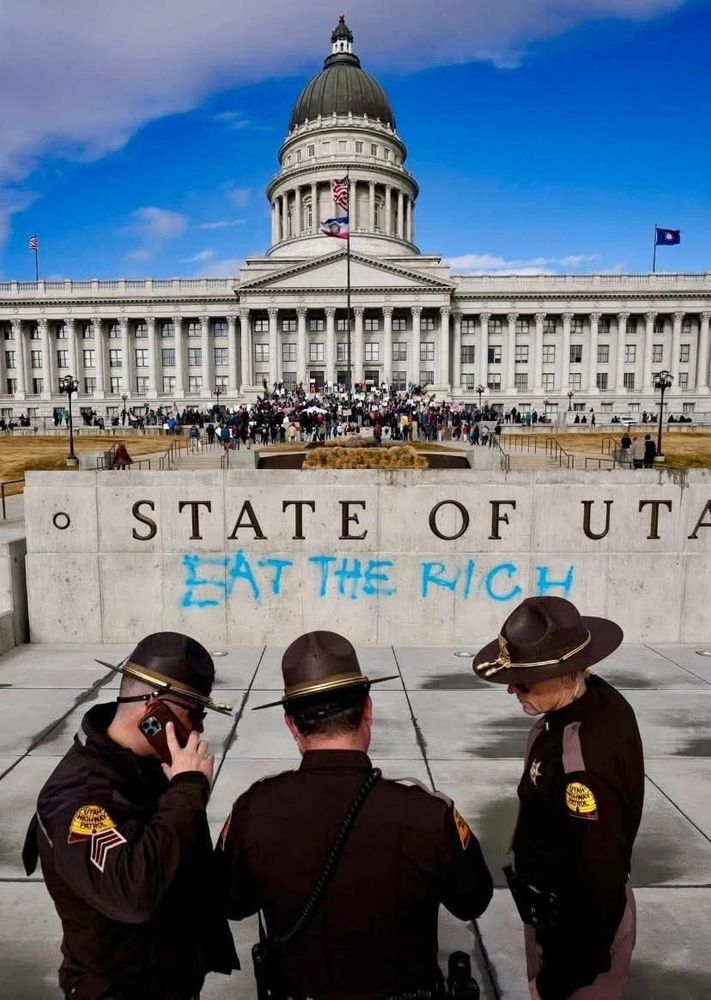 utah state capitol in the background with a gathering of people
state of utah sign with EAT THE RICH spray painted in blue
3 cops stand around huddled up, one on the phone in front of the sign
