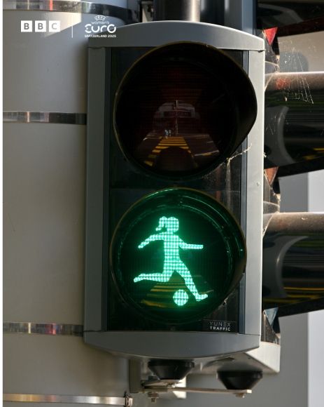 Traffic lights on green where the usual "green man" has been replaced with a woman kicking a football. 