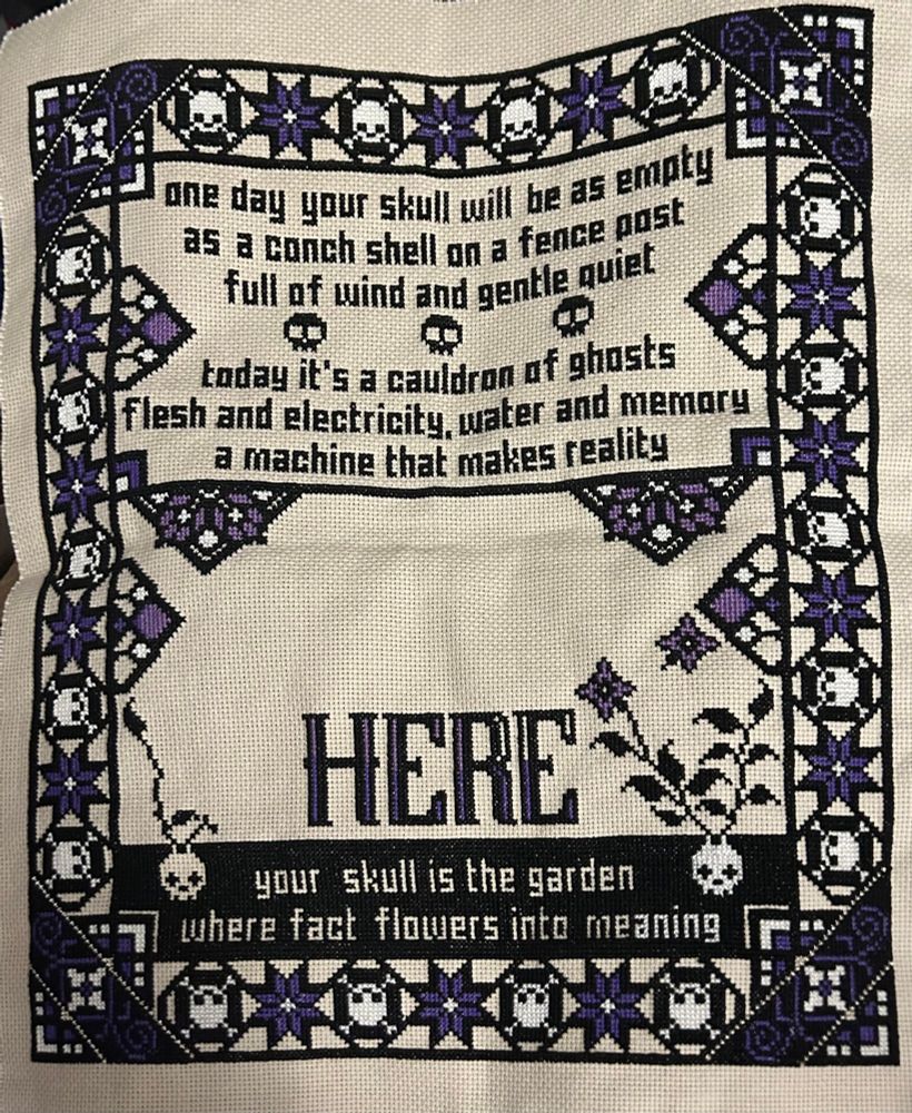 A frame of eight-pointed purple stars and white skulls are cross stitched around words that say, so far "one day your skull will be as empty / as a conch shell on a fence post / full of wind and gentle quiet / today it's a cauldron of ghosts / flesh and electricity, water and memory / a machine that makes reality". At the bottom: "Here your skull is the garden where facts flower into meaning."