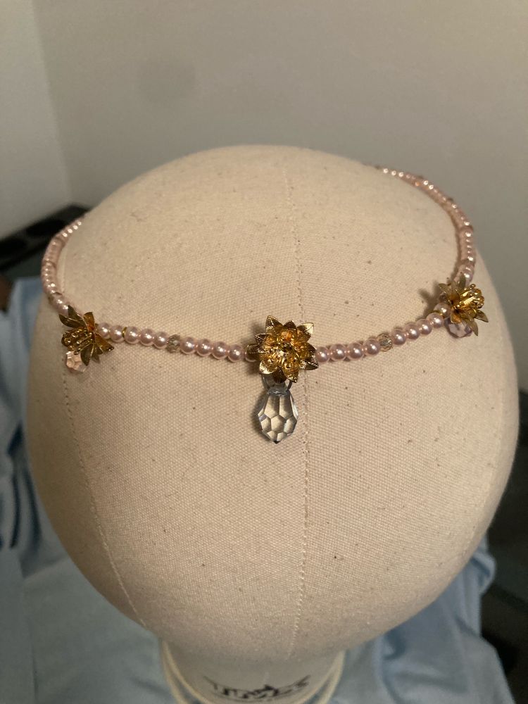 A small pink pearl circlet with 3 gold flowers spaced evenly across the front, each with a dangling crystal drop. It is displayed on a plain wig head