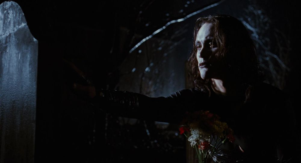 The Crow, Eric Draven, holds some flowers and sadly touches the headstone of his fiancée.