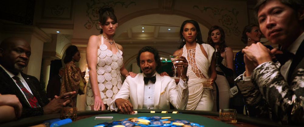 Samba Schutte's "Pluribus" character Mr. Diabaté at a casino table dressed in a white tux with one woman at each shoulder. #OFMD #BondJamesBond