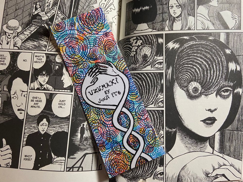 handdrawn bookmark laying on the open pages of uzumaki by junji ito. the bookmark features a pair of intertwining arms twisting around each other to reach for each others hand. behind them are countless colorful spirals overlapping in yellow, blue, red, green, and purple. 