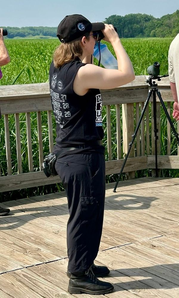 person looking through binoculars at something off camera. they are wearing all black and a sleeveless t shirt. 