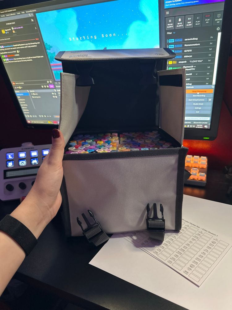 there is a bag of ohuhu alcohol markers on a desk, along with a swatch sheet. there is a monitor in the background with obs (a streaming program) open with a “starting soon” screen open