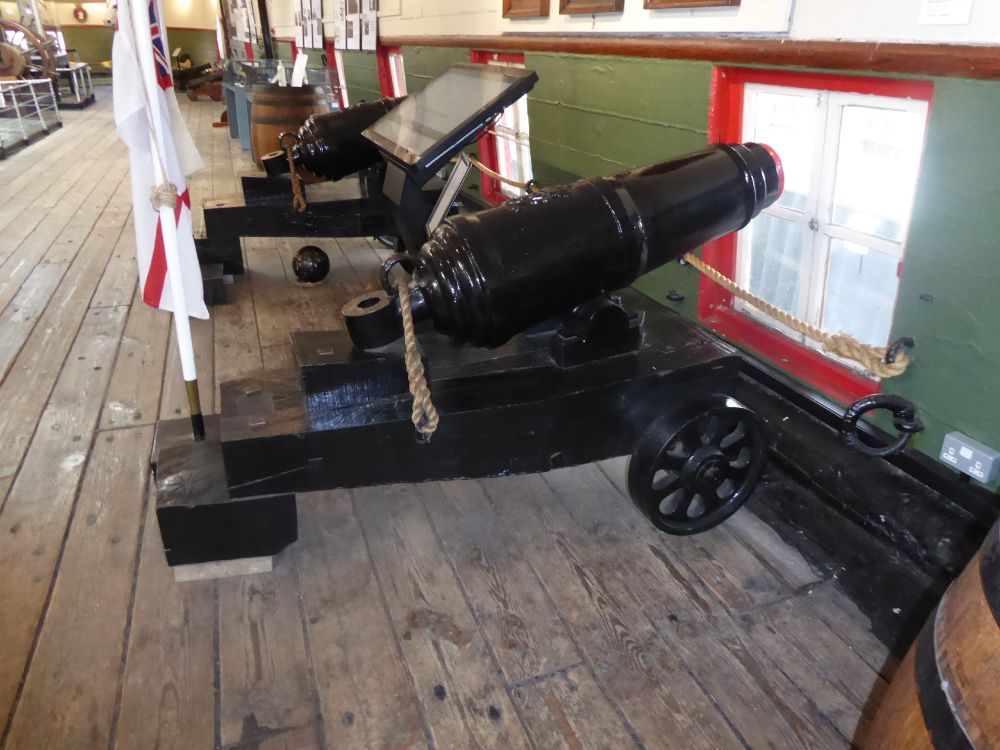 This is a late-model carronade mounted on a battery carriage (two iron wheels, which would damage the deck if you fired it on a wooden vessel). At sea, carronades were mounted on slides, rather than carriages. It is much shorter than a gun, with an elevation screw at the rear, rather than a quoin (which was a wooden wedge to control elevation). The elevation screw is missing, but the loop for the screw is visible behind the breech of the gun. There is a breeching loop above the button. Two reinforces - the first with a royal cipher (George the Third in this case). The trunnions are below the barrel, as on all carronades. Red tompion in the muzzle. There is a second carronade behind. Both are on the port side of the weather deck of HMS Unicorn.