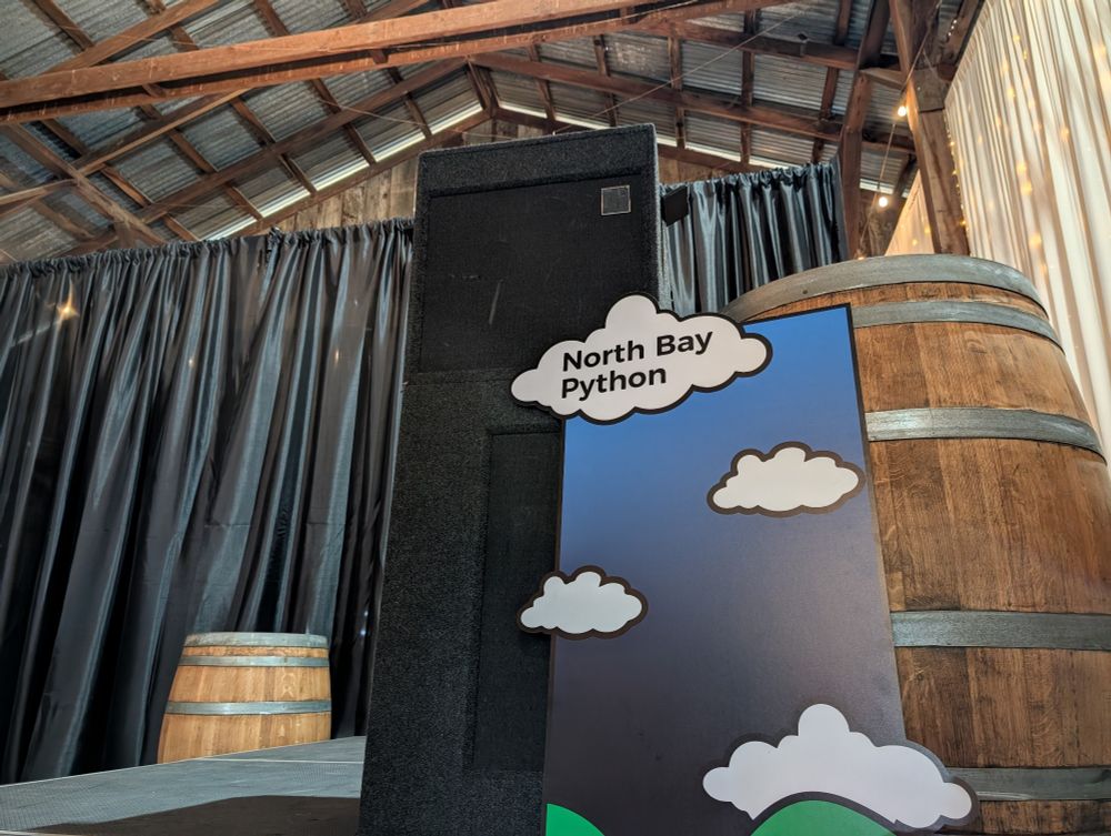 A photo of the lectern front featuring the green hills, blue skies, and fluffy white clouds of the North Bay Python branding leaning against a wine barrel on stage.