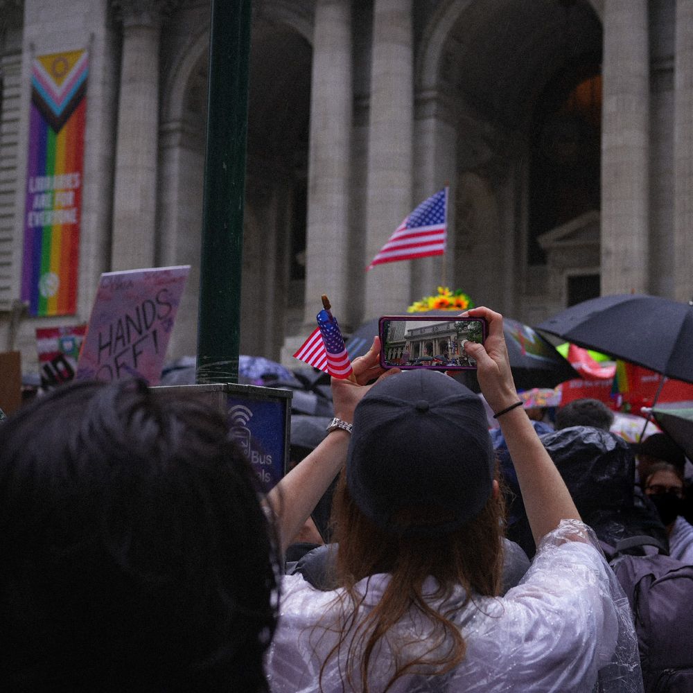 a collection of damp protestors in front of the main NY public library branch. the focus of the image is a person taking a photo on their phone of a US flag in front of the library. a large pride banner hangs on the wall of the library reading 'libraries are for everyone'