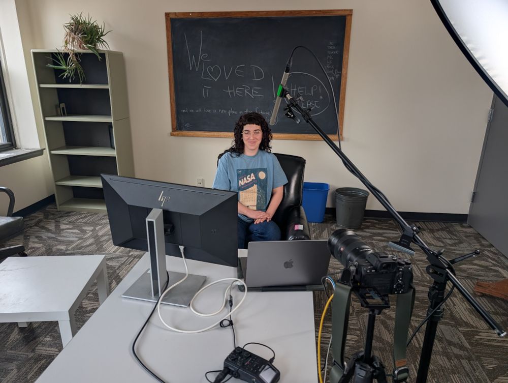 Casey Ivanovich sits in front of a live streaming setup in a nearly empty NASA GISS office, preparing to give a live talk. 
She's wearing a NASA GISS t-shirt