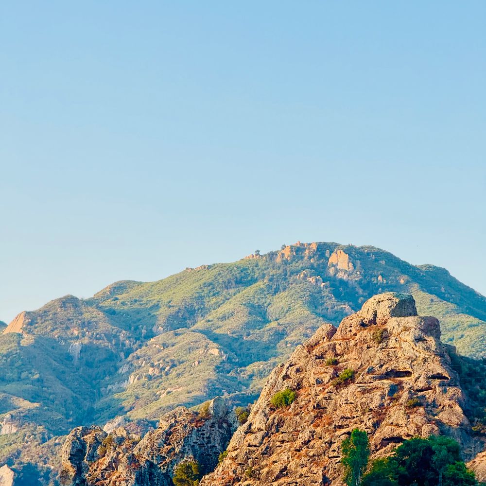 Blue skies and green hills in the background with rocky hills with small caves in foreground. The cave openings look like eyes and mouths to me, so I see faces. Thus the comment about stories to tell. 