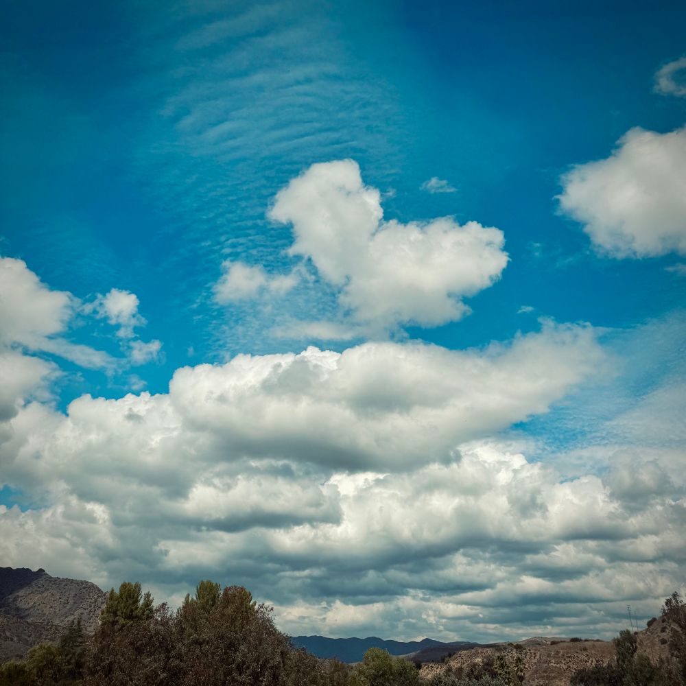 Vivid blue sky with big fluffy white and gray clouds rolling through. Hills and trees across the bottom. 