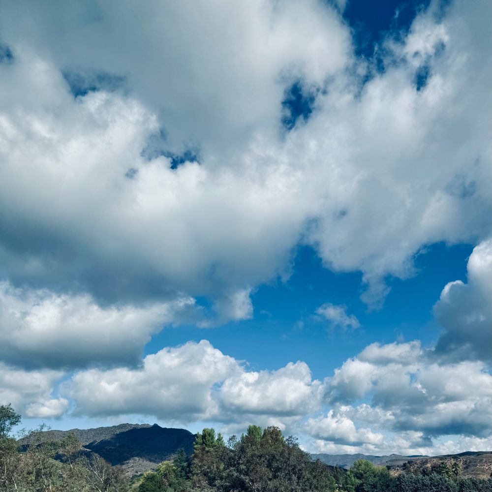 Puffy gray and white clouds fill the sky but areas of bright blue shine through. A distant hill is halfway shadowed by the cloud above with sun hitting the green treetops across the bottom of the photo. More hills are seen in back along the horizon. 