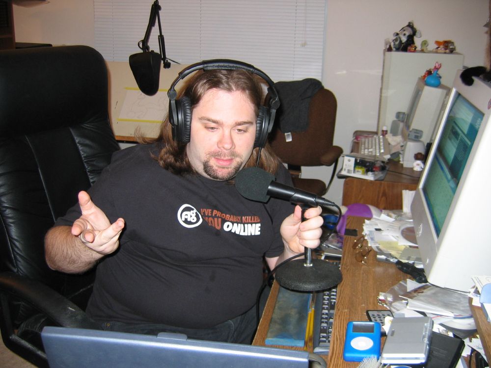 A picture dated in January of 2006 of me doing my podcast. A CRT monitor with Winamp and sound mixers on it, a Shure C608 mic on a table stand being held, an iPod Classic and Nintendo DS on the messy desk, and me with a very early goatee (I just started growing it) wearing an All Games Radio shirt, where my show originally was broadcast.