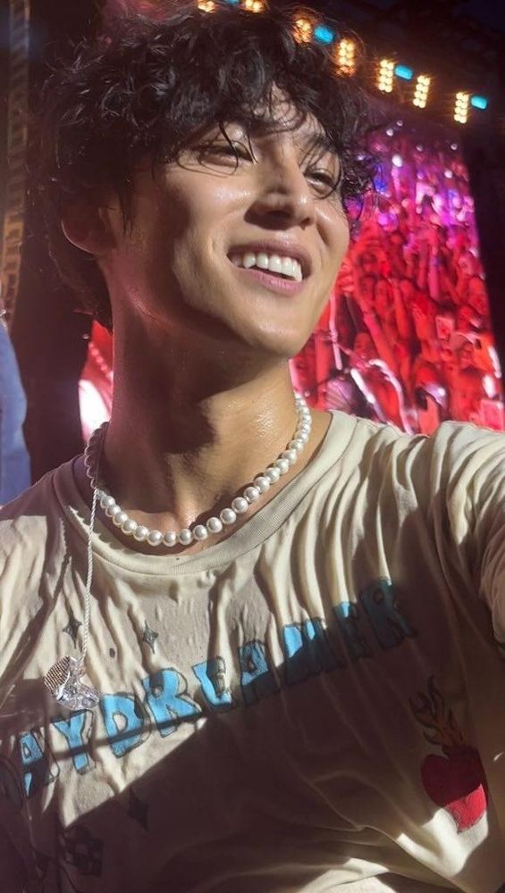 A picture of Mingyu from the chest up on stage at a concert. He is smiling. His shirt is wet and sticking to him, maybe sweat maybe someone poured water on him. His hair is curly and he is wearing a short pearl necklace.