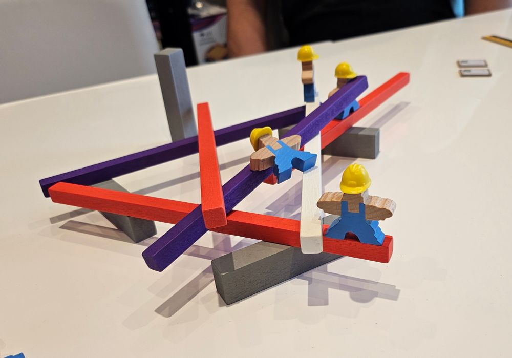 A game of Men at Work, three wooden workers on a mess of girders, one is almost hanging in the air, barely touching two planks. 