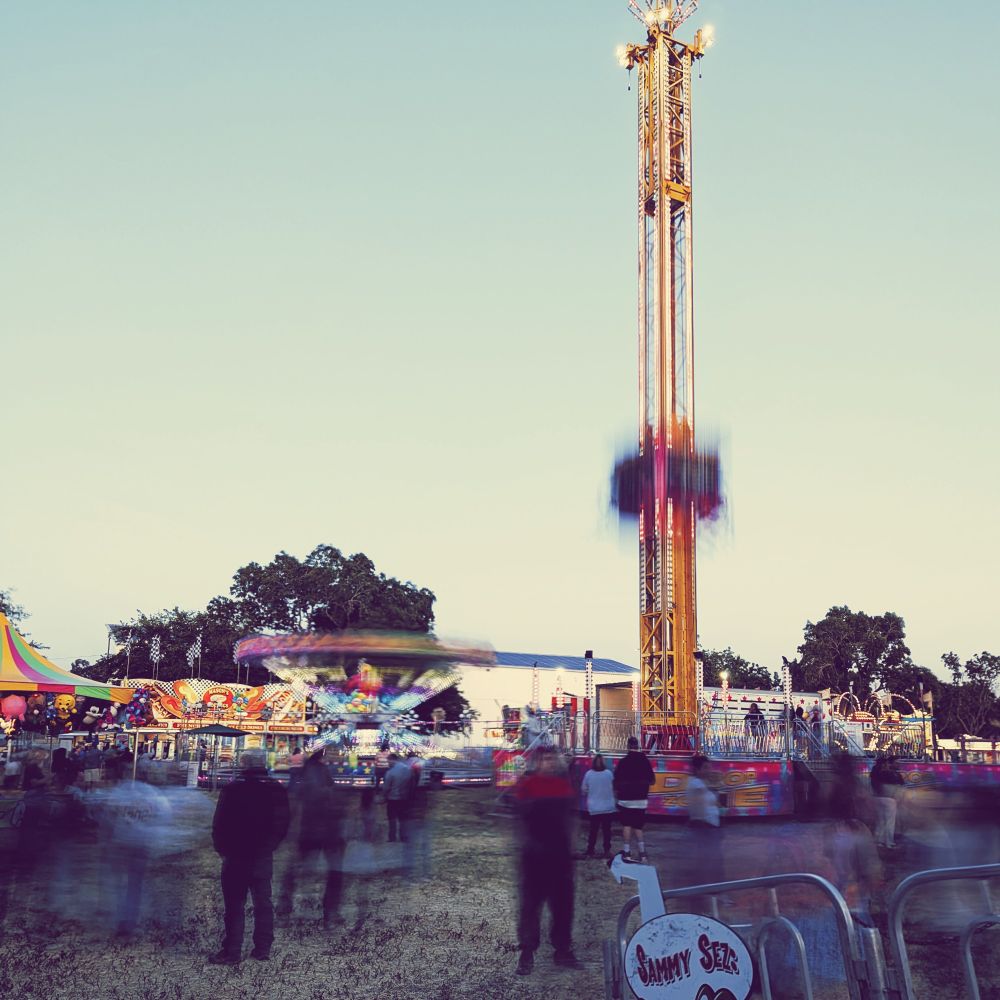 Drop Tower at the fair; riders are seated and secured on a little elevator and brought to the top, then experience a brief moment of free fall until they are halted and released. Crowds in the foreground are blurred into illegibility.
