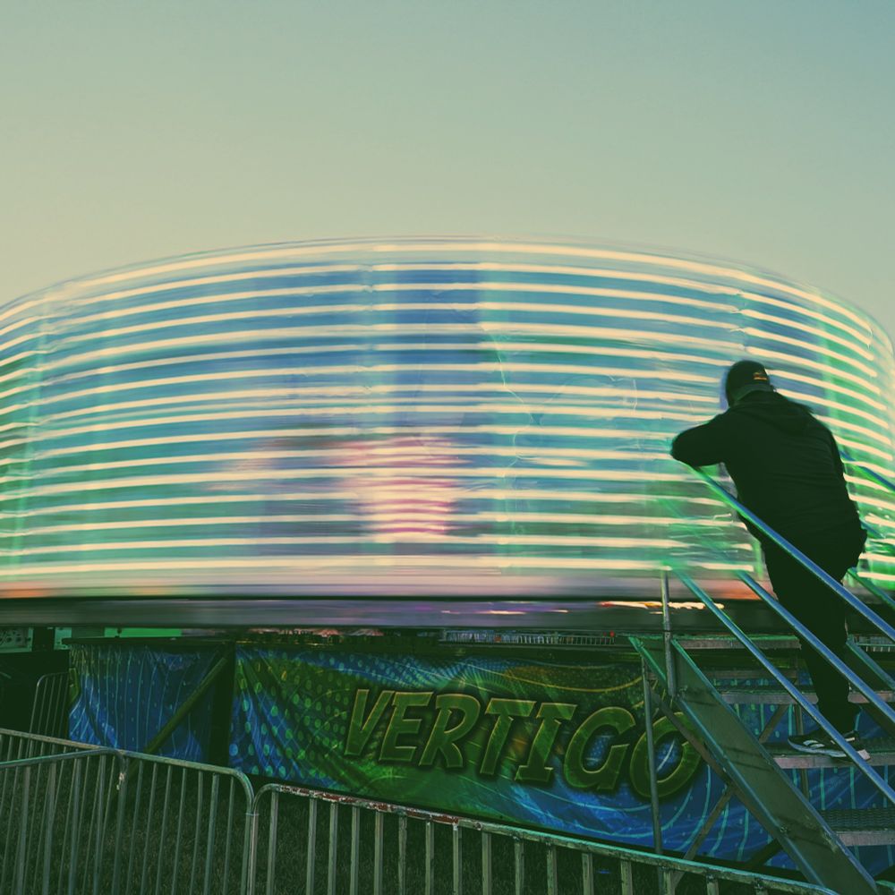 Ride operator standing on the boarding stairs for Vertigo, an open ride that lines riders along the perimeter of a cylindrical platform, then spins to press them against the walls. By this time the ride is going fast enough to blur the lights into solid lines.