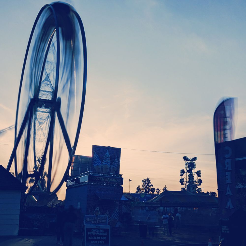 Ferris Wheel at Contra Costa County Fair at sunset; a long exposure has been used to blur the wheel's motion on the left, echoed by a flapping banner on the right advertising ice cream. Other booths, people, and rides are rendered as stark silhouettes, having exposed for the sky.
