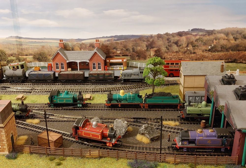 A model railway scene with six colourful steam engines arranged around a yard. There is a station in the background with a small diesel engine pulling stone wagons.