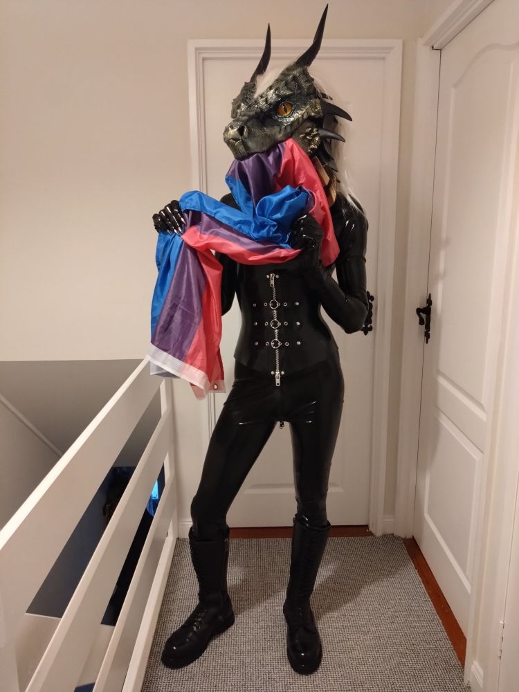 A fursuiter wears a green dragon head with white hair and yellow eyes, a black rubber catsuit and corset with leather knee high boots and black pvc claw gloves. He is standing in a hallway, eating a bi pride flag