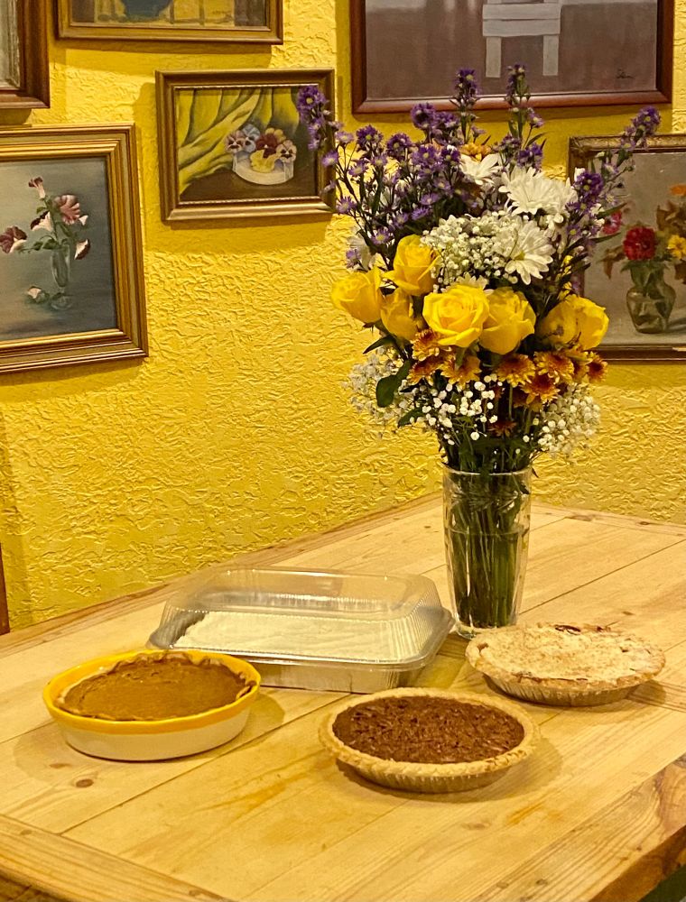 A pumpkin pie, a pecan pie, an apple pie & a spice cake with a gorgeous vase of yellow roses, daisies, & mums. 