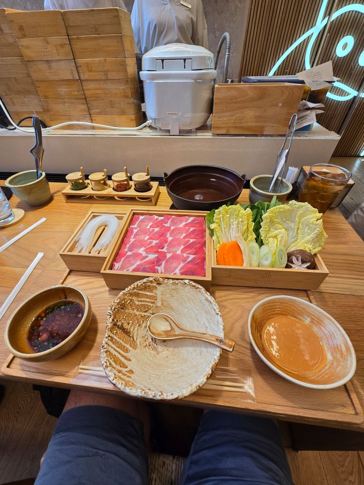 The image captures a beautifully arranged table set for a meal, likely in a cozy restaurant setting. The table itself is wooden, exuding a warm and inviting atmosphere. At the center of the spread is a bamboo tray, meticulously filled with thinly sliced raw meat, likely beef or pork, arranged in a neat rectangular pattern with each slice evenly layered.

Beside it, another wooden tray holds a vibrant selection of fresh vegetables, including crisp, light-green napa cabbage leaves, bright orange carrot slices, earthy brown shiitake mushrooms with their characteristic umbrella-like caps, delicate clusters of white enoki mushrooms, and freshly cut green onions.

Two bowls are positioned close by—one containing a thick, sesame sauce and the other holding a ponzu sauce speckled with chopped herbs and spices. A wooden spoon rests inside one of the bowls, ready for serving. Nearby, a small container holds a pair of tongs, likely for handling the raw ingredients.

Also present is a glass filled with roastes tea, its amber color complemented by condensation on the outside, hinting at its refreshing coolness. In addition, a set of condiments sits neatly in small wooden containers with lids, adding variety and customization to the meal.

In the background, a rice cooker and a stack of wooden boxes. The setting, with its communal layout and fresh, high-quality ingredients, strongly suggests a traditional hot pot or shabu-shabu dining experience, where individuals cook their selected meats and vegetables in a bubbling broth.

The composition of the image exudes a warm, shared dinings to additional ingredients experience with an emphasis on fresh flavors, interaction, and tradition. The visual arrangement, colors, and textures make it a feast for the senses.
