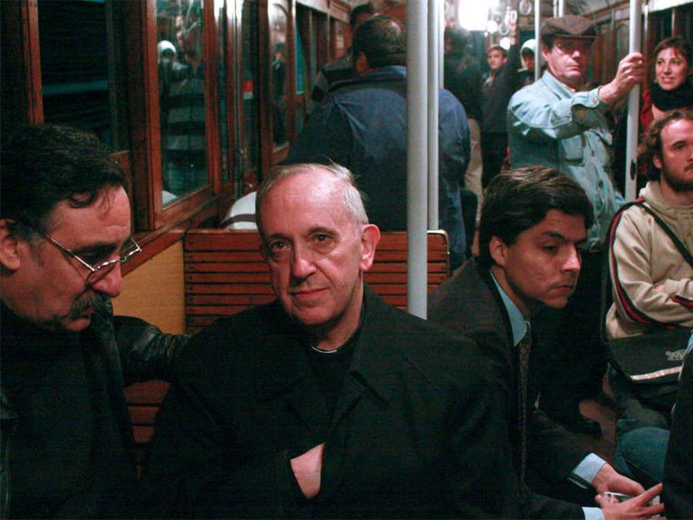 
foto do papa francisco, antes do papado, sentado em um vagão de metrô cheio de pessoas, em buenos aires. ele está vestido de roupas pretas, agasalhado, e encara as lentes da câmera com um olhar calmo e profundo.
