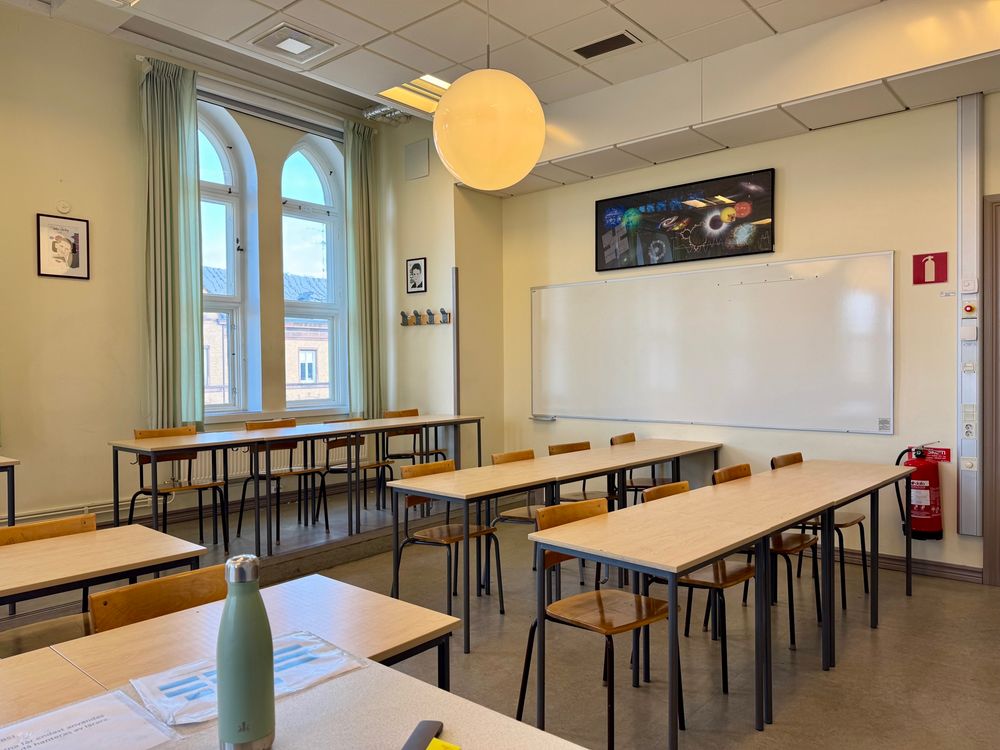 Bilden visar höger sida av ett klassrum. I förgrunden syns delar av en kateder och några bänkrader. Sedan syns 3 bänkrader med 4 bänkar i varje rad. På väggen vid sidan av bänkraderna finns en whiteboardtavla. På väggen bakom bänkraderna finns två stora fönster med ljusgröna gardiner. 