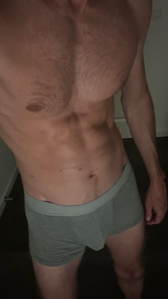 A gay man standing in low light wearing tight grey briefs. His toned, hairy torso and relaxed stance are visible in the soft darkness. The lighting and pose reflect an intimate, confident gay top energy winding down for the night.