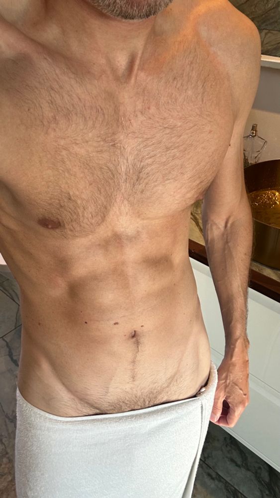 A gay man stands in a bathroom with a towel loosely wrapped around his hips, showing off a toned, hairy chest and defined abs. His hand grips the towel, hinting at what’s underneath. The lighting is natural, suggesting a confident, flirty gay top getting ready to start his day.