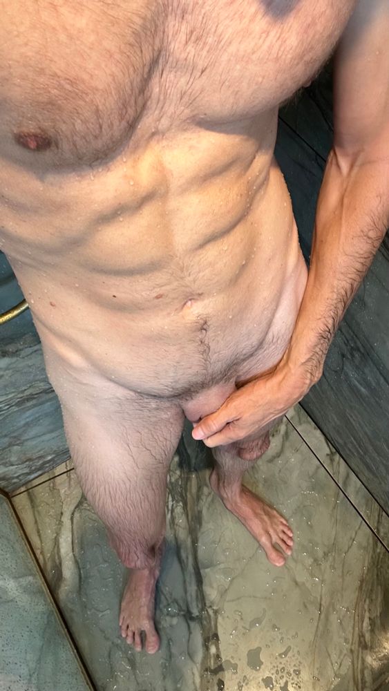 A gay man standing nude in a wet shower, his toned, hairy torso and cock partially covered by his hand. Water droplets still glisten on his skin, and the photo is shot from above to capture his entire stance on the tiled floor. His pose and teasing grip radiate confident, dominant gay top energy just after rinsing off.